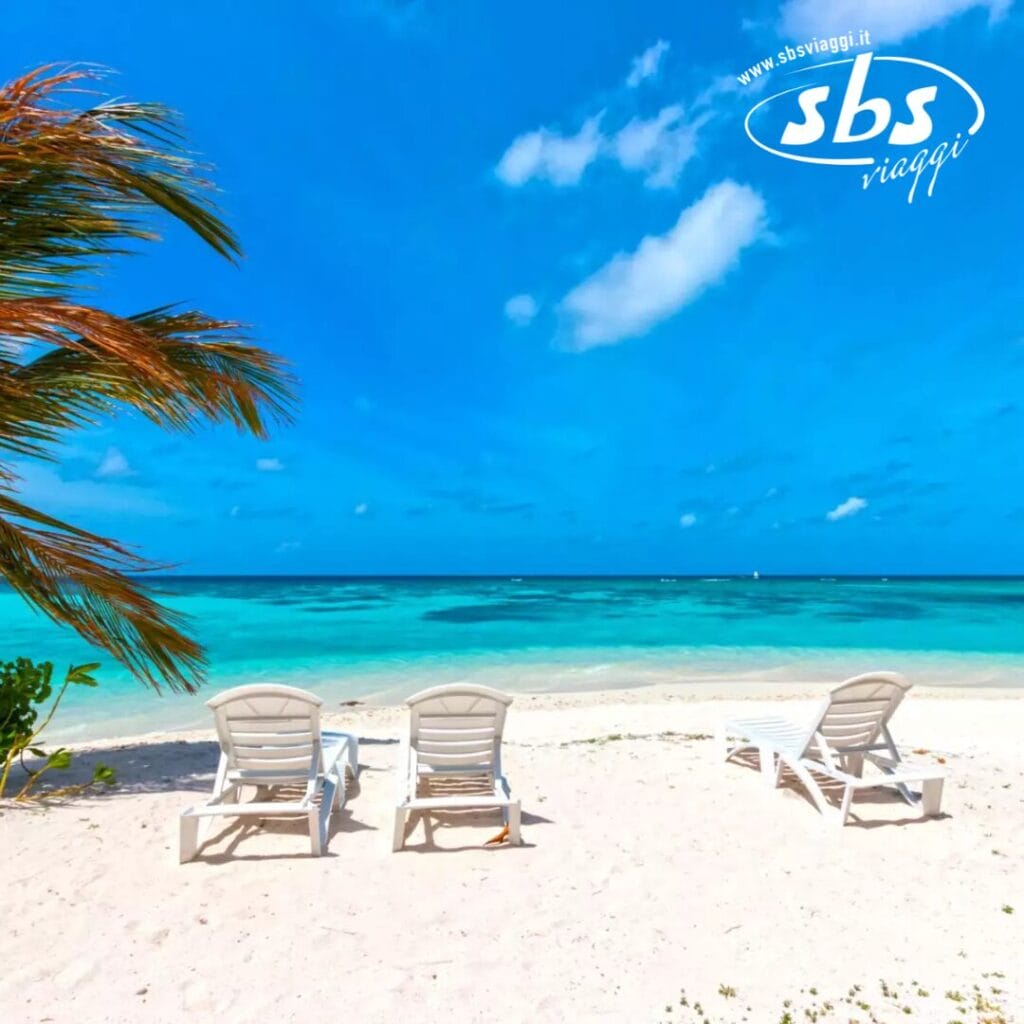 Tre sedie a sdraio vuote su una spiaggia sabbiosa, parte dell'esclusiva esperienza del Plumeria Hotel, si affacciano sull'oceano turchese sotto un cielo azzurro e limpido, con foglie di palma che ondeggiano dolcemente sulla sinistra.