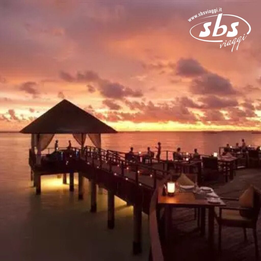 Godetevi un tramonto indimenticabile presso il ristorante sull'acqua del Sun Siyam Iru Fushi alle Maldive, dove luci brillanti illuminano il ponte di legno sopra un oceano calmo.