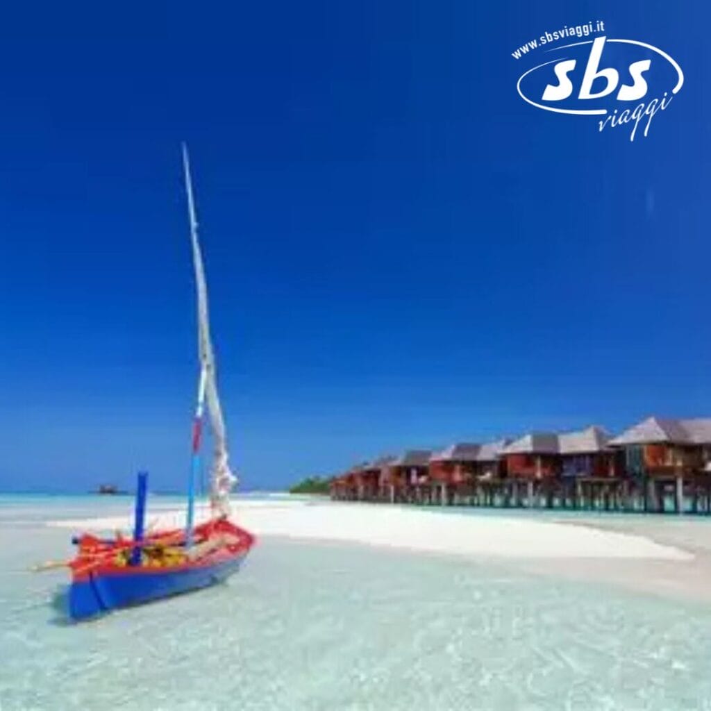 Una barca a vela colorata abbellisce la spiaggia limpida e poco profonda, con bungalow sull'acqua che si stagliano contro un cielo azzurro brillante. L'inconfondibile logo