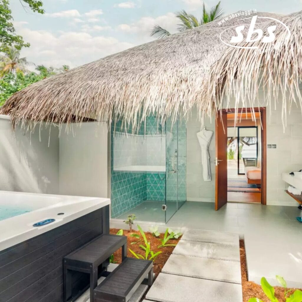 Spa tropicale all'aperto con tetto di paglia, doccia con vetrate e jacuzzi accanto a una porta che conduce all'interno. Concedetevi il relax in questa oasi di Nova Maldives. Un accappatoio è appeso all'ingresso, invitandovi a rilassarvi con stile sull'isola di Vakarufalhi.