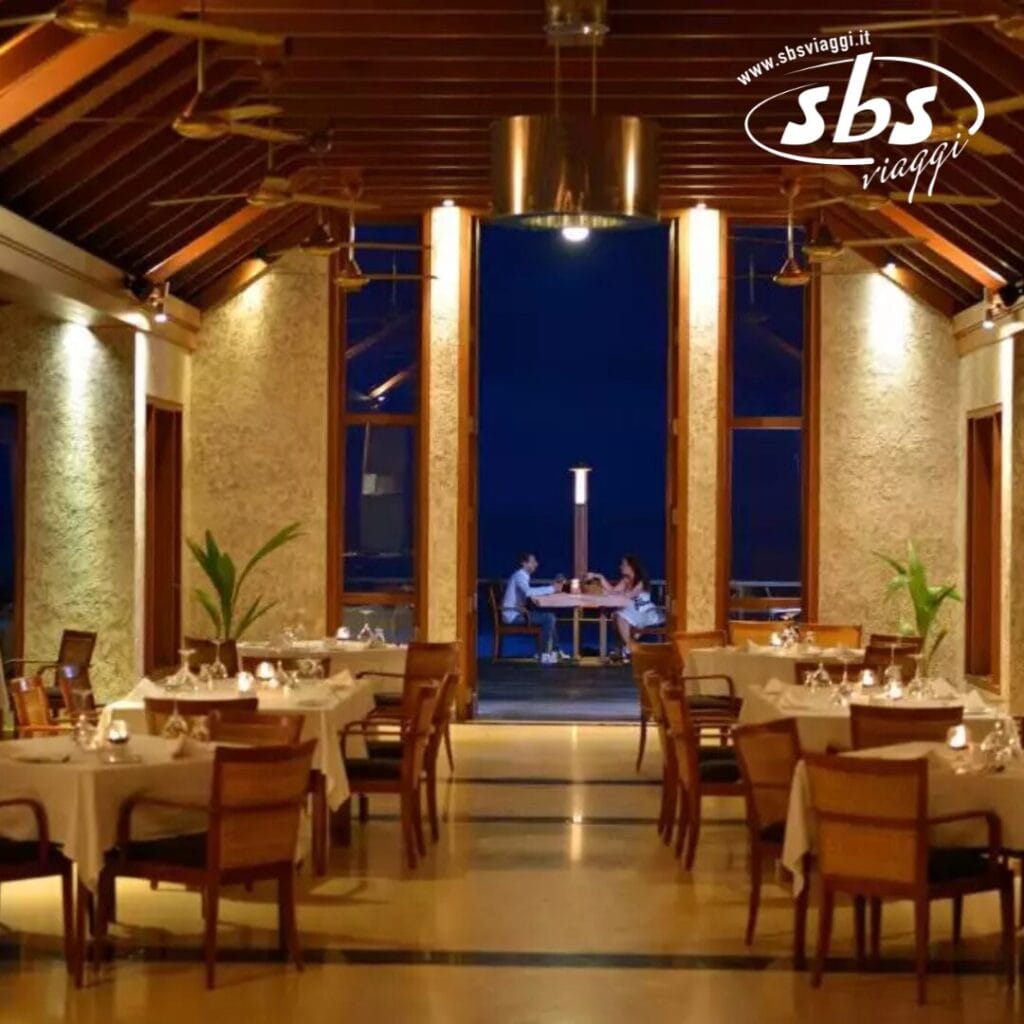 Un elegante ristorante interno con soffitto in legno e tavoli apparecchiati. Due persone siedono a un tavolo vicino alle porte aperte, godendosi la bozza automatica di un incantevole cielo serale sullo sfondo di un'atmosfera soffusa e illuminata.