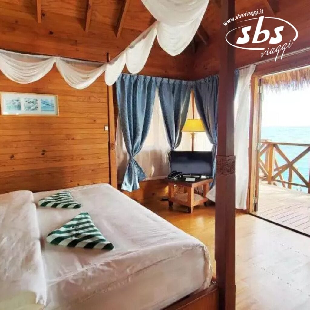 Una camera da letto accogliente in legno con un letto grande, asciugamani a strisce verdi e bianche, una piccola area soggiorno e tende che si aprono su una vista sull'oceano a Fihalhohi. Il logo