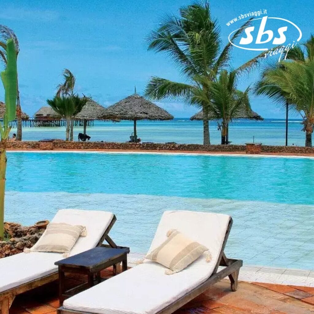 Due sedie a sdraio bianche a bordo piscina presso l'Igv Dongwe Club offrono un rifugio perfetto, circondati da palme e ombrelloni di paglia, con una vista mozzafiato sull'oceano sotto un cielo azzurro e limpido.