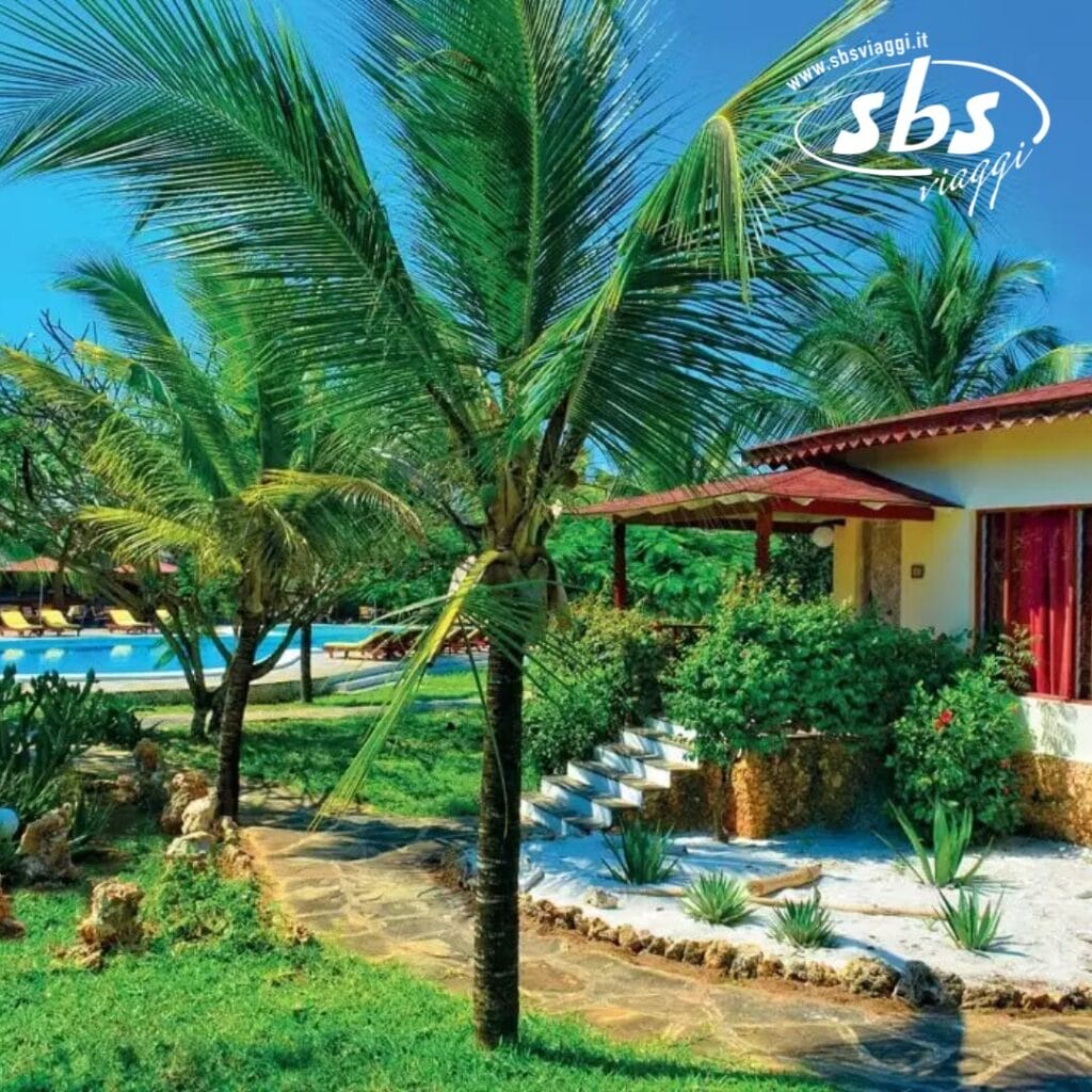 Giardino tropicale con palme e un sentiero in pietra che conduce a una casa. Una piscina è visibile sullo sfondo sotto il cielo azzurro e terso di Crystal Bay, che riflette l'atmosfera serena che ricorda il lusso distintivo di Veraclub.