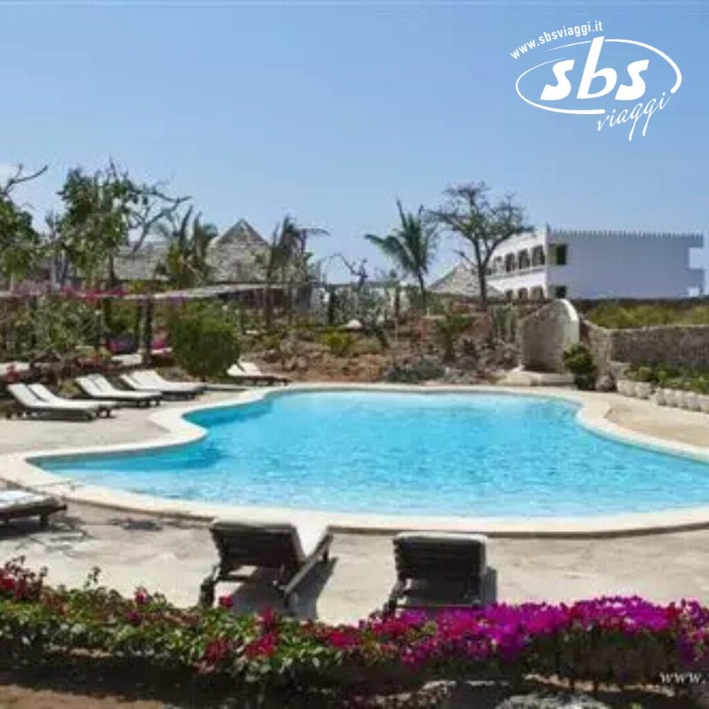 Il Jumbo Watamu Resort dispone di una piscina all'aperto circondata da sedie a sdraio e vegetazione lussureggiante, con un immacolato edificio bianco sullo sfondo sotto un limpido cielo azzurro.
