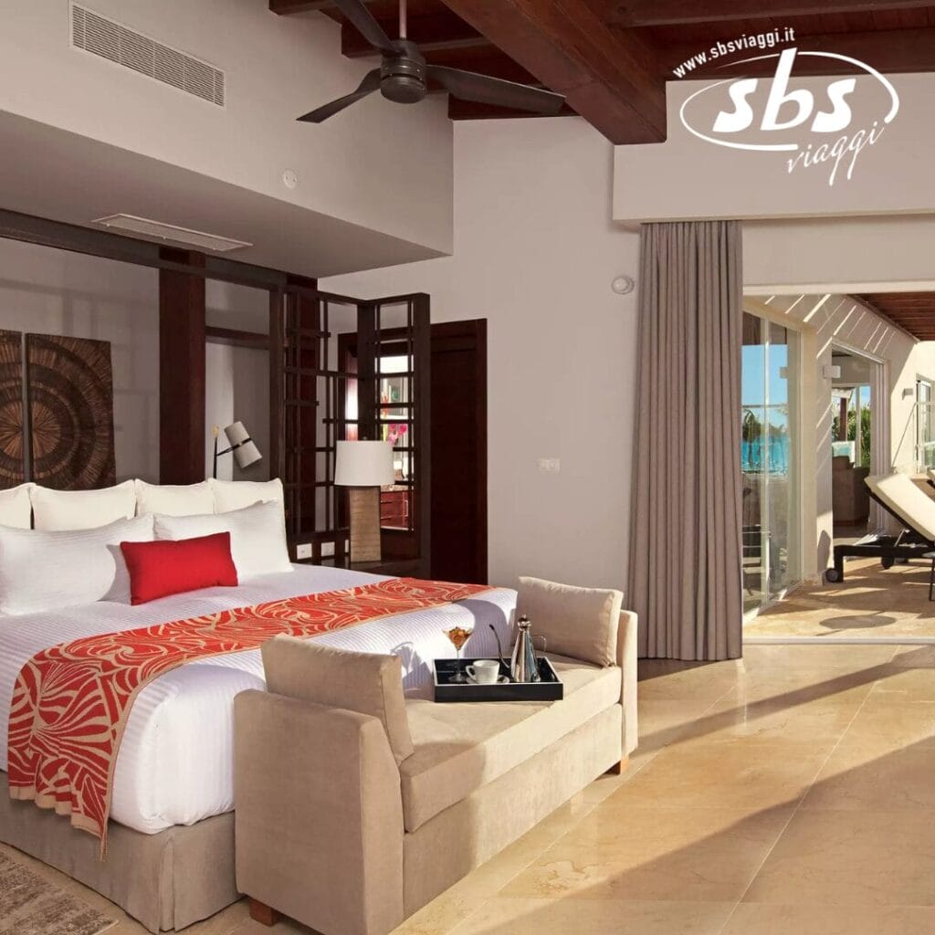 Lussuosa camera d'albergo con letto king size, accenti rossi, un piccolo divano ai piedi e un balcone aperto con vista su La Romana. Vivi i sogni in stile Seaclub mentre ti godi la vista tranquilla. Logo