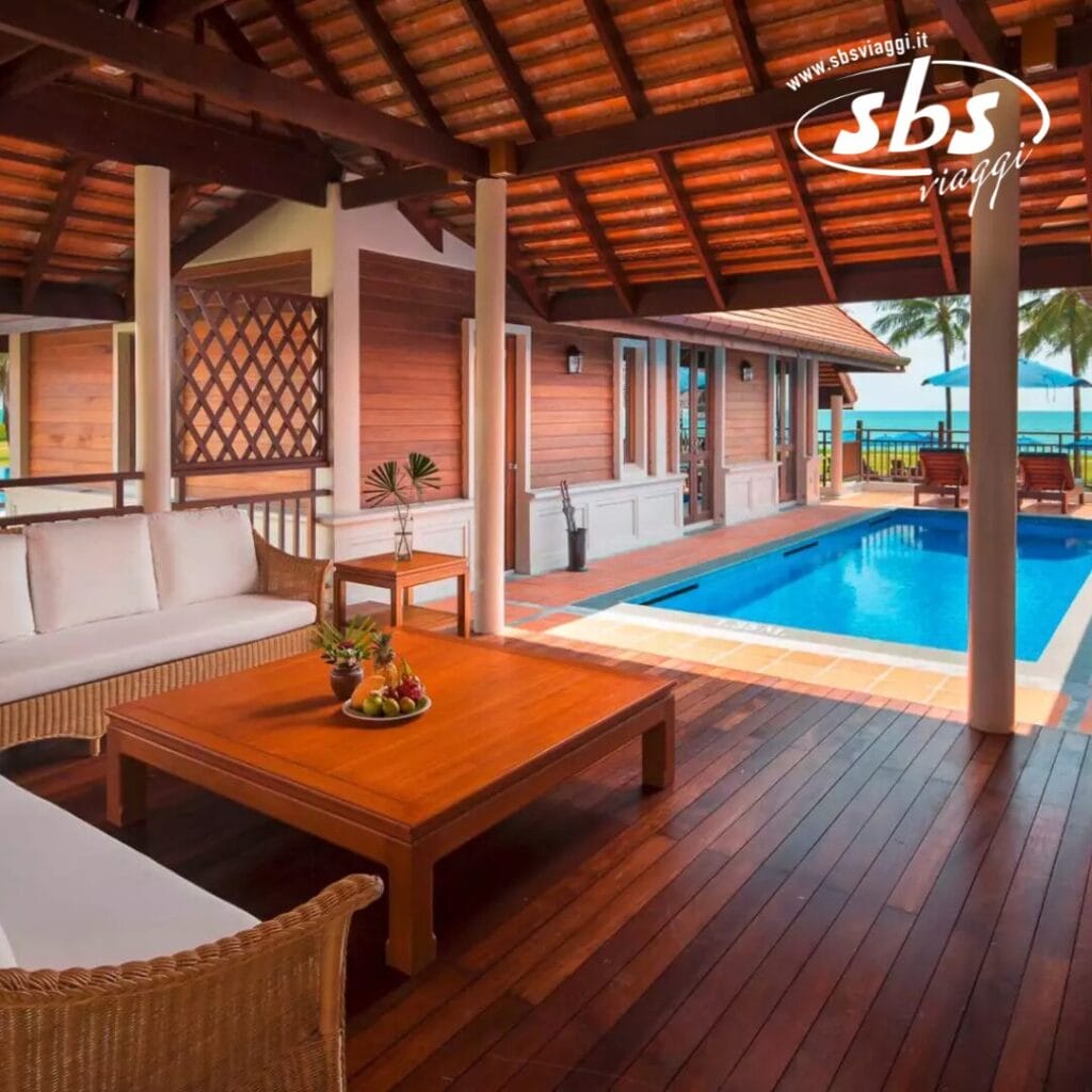 Lussuoso patio esterno con terrazza in legno, divano, tavolino e fruttiera, con vista sulla piscina e sull'oceano sotto un tetto incantevole.