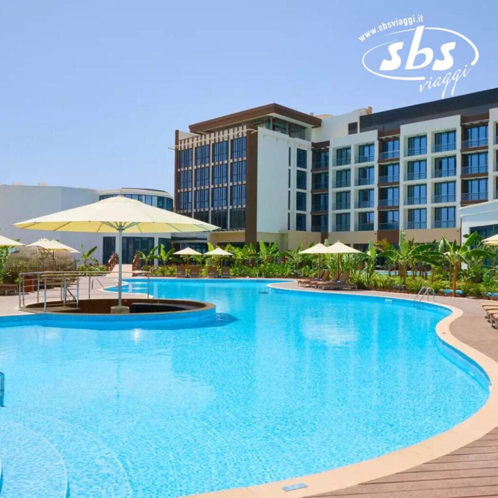 L'area piscina esterna dell'hotel è caratterizzata da acqua cristallina e azzurra, circondata da sedie a sdraio e ombrelloni. Un edificio moderno si erge sullo sfondo in mezzo a una vegetazione lussureggiante, sotto un cielo terso e soleggiato.