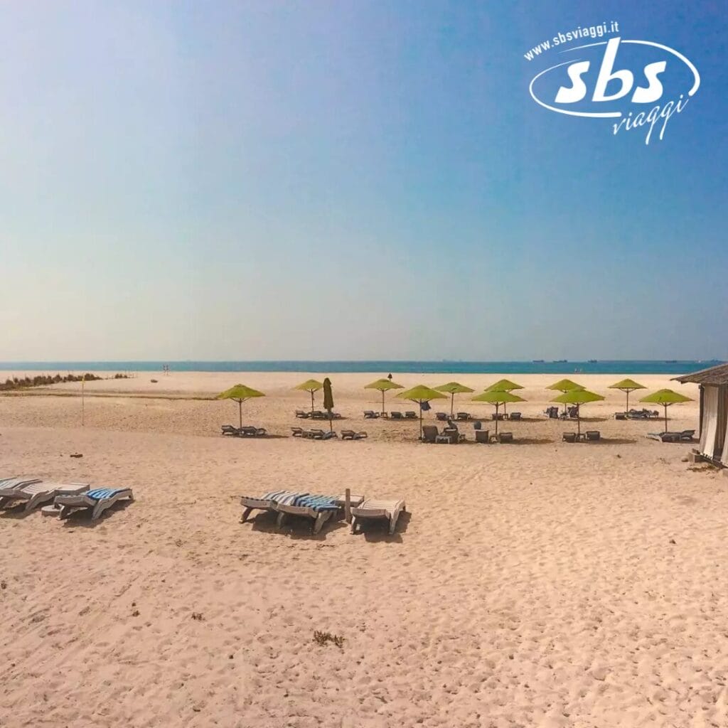 La tranquilla spiaggia sabbiosa dell'Hilton Resort di Salalah è attrezzata con numerose sedie a sdraio e ombrelloni verdi, tutti perfettamente allineati verso l'oceano sotto un cielo azzurro e limpido.