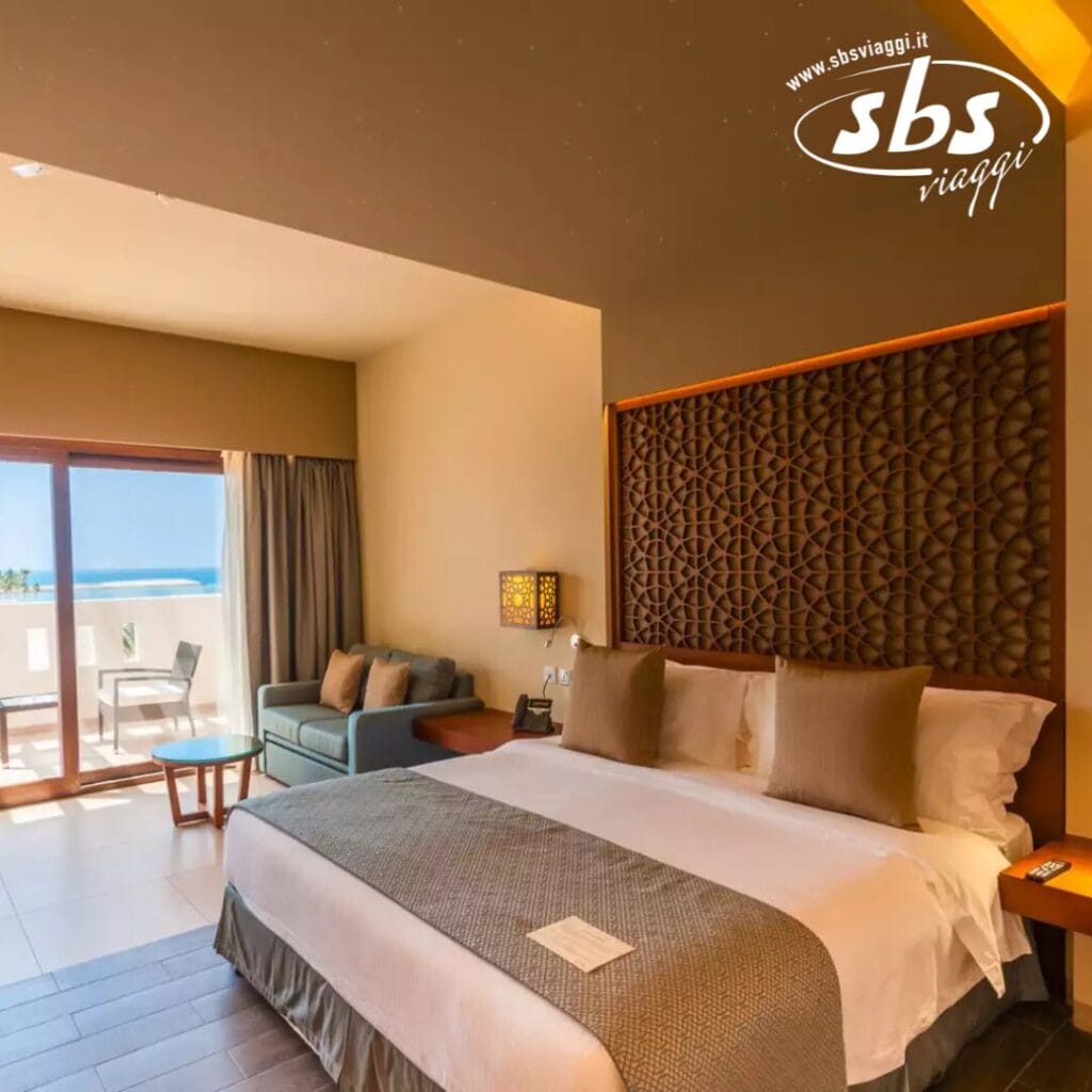 La camera dell'hotel Seaclub Fanar è dotata di un letto grande con decorazioni marroni e beige, un comodo divano e un balcone che offre una vista mozzafiato sull'oceano. Sul balcone troverete un tavolino e una sedia. Il logo