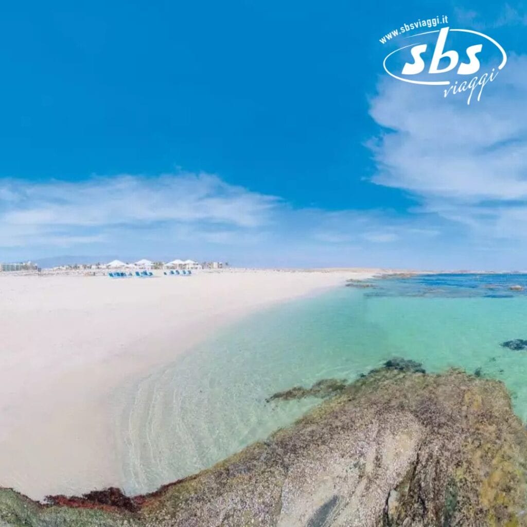 Spiaggia sabbiosa con acqua azzurra e cristallina e uno sfondo roccioso. Sedie a sdraio e ombrelloni sono visibili in lontananza sotto un cielo parzialmente nuvoloso, creando una scena pittoresca che ricorda una bozza automatica. Il logo