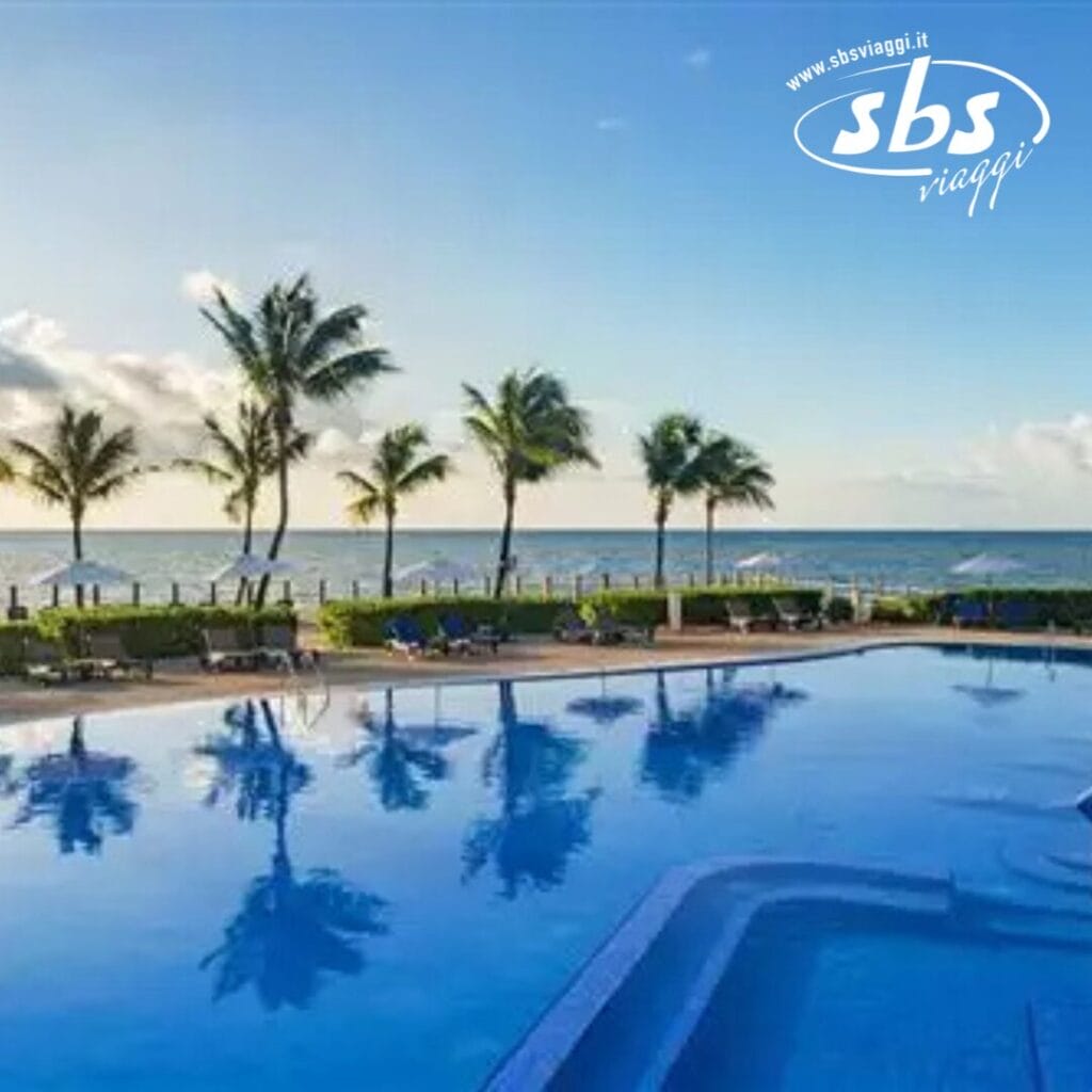 Una piscina serena sulla spiaggia con palme e una vista mozzafiato sull'oceano all'alba, catturando perfettamente la tranquillità. Il logo