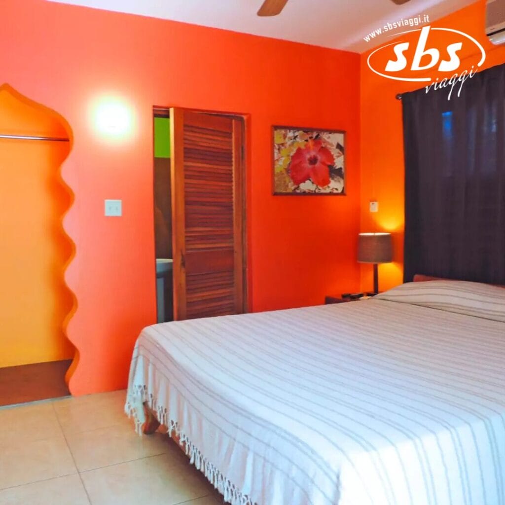 L'atmosfera del Sunrise Club riempie questa camera da letto colorata, con una parete arancione, un comodo letto matrimoniale, una lampada elegante e un dipinto incorniciato. Una porta conduce a un'altra stanza mentre una tenda copre la finestra. Il logo