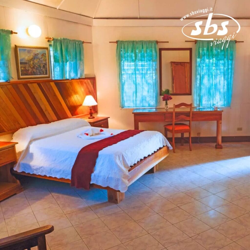 Una camera d'albergo accogliente al Samsara Beach Resort è dotata di un grande letto con testiera in legno e biancheria bianca impreziosita da un runner rosso. Due finestre con tende color verde acqua lasciano entrare la luce naturale, mentre una scrivania e una sedia sono sistemate in modo invitante nell'angolo.