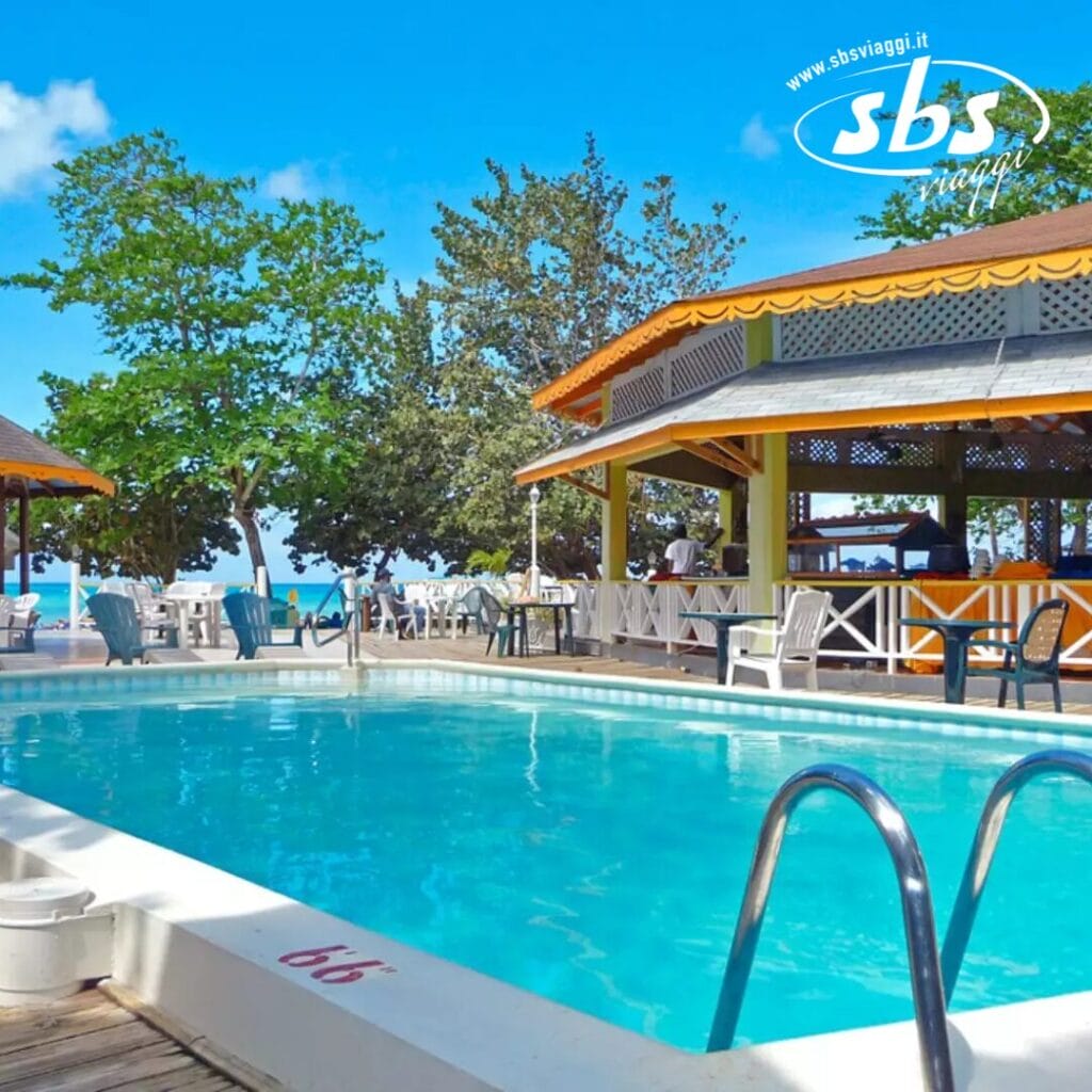 La piscina esterna del Merril's Beach Resort è dotata di una scala di metallo, circondata da tavoli e sedie. Alberi e un edificio con un tetto giallo esaltano lo scenario, mentre il mare blu luccica in lontananza.