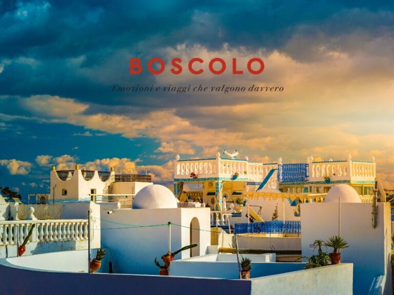 Edifici bianchi in stile mediterraneo con accenti blu sotto un cielo spettacolare, che evocano il fascino di Hammamet. La scritta "Boscolo" sulla scena.