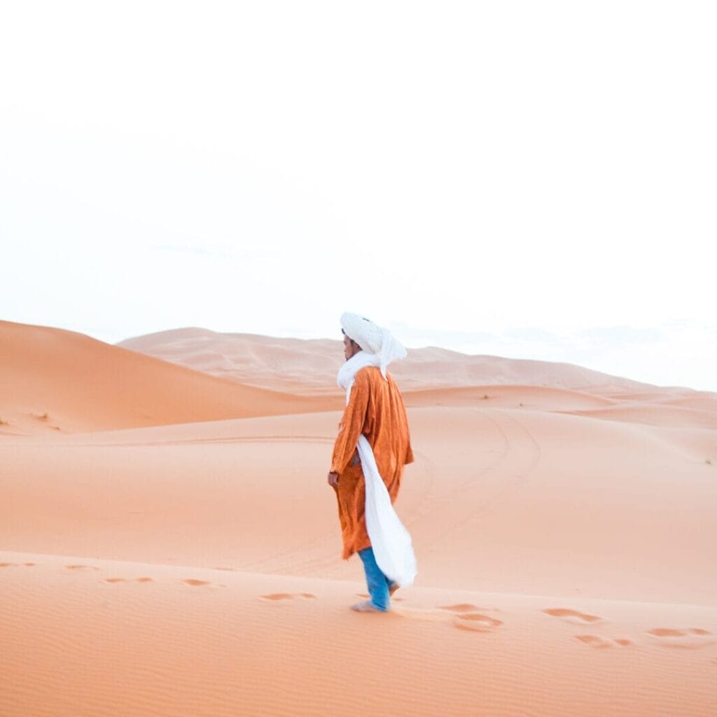 Una persona in abiti tradizionali intraprende un tour affascinante, camminando tra le vaste dune di sabbia del Sahara sotto un cielo limpido, che ricorda i viaggi che partono da Marrakech.