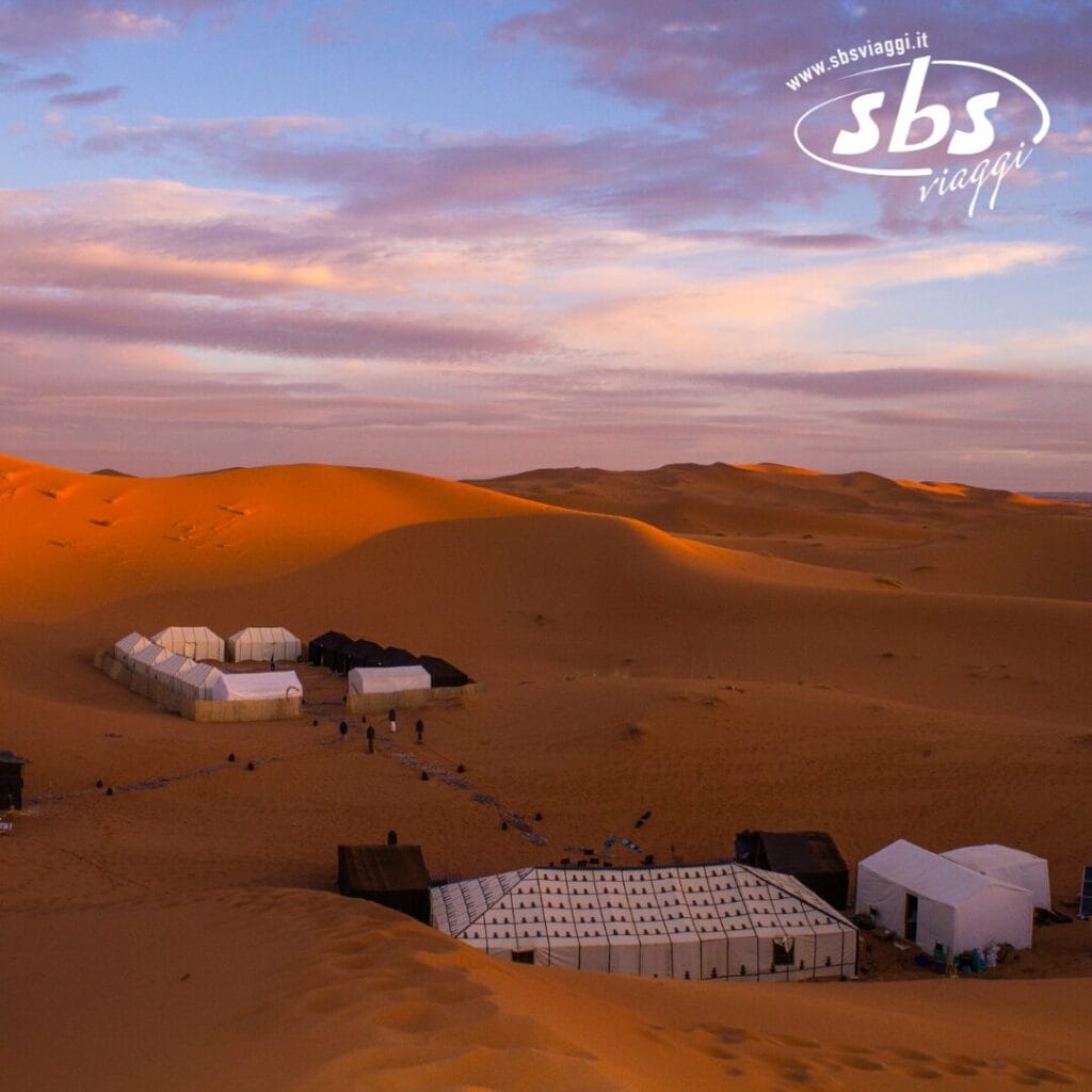 Tende sulle dune sabbiose del Sahara sotto un cielo nuvoloso al tramonto, che offrono uno sguardo sereno nel cuore dell'avventura. Logo