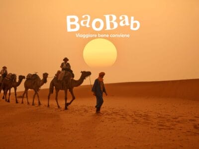 Persone con cammelli camminano nel deserto al tramonto, avvolte nella magia del Sahara. Il testo recita: "Baobab, viaggiare bene conviene".