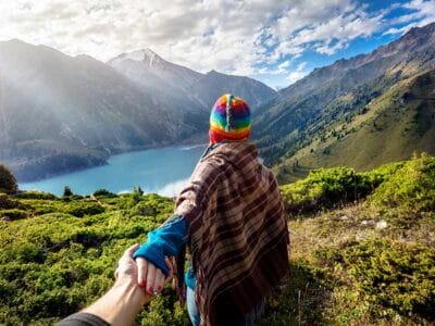 Una persona con un cappello colorato tiene la mano di un'altra mentre ammira un lago di montagna, circondato da verdi colline e un cielo parzialmente nuvoloso, come se fosse stata immortalata da una bozza automatica.