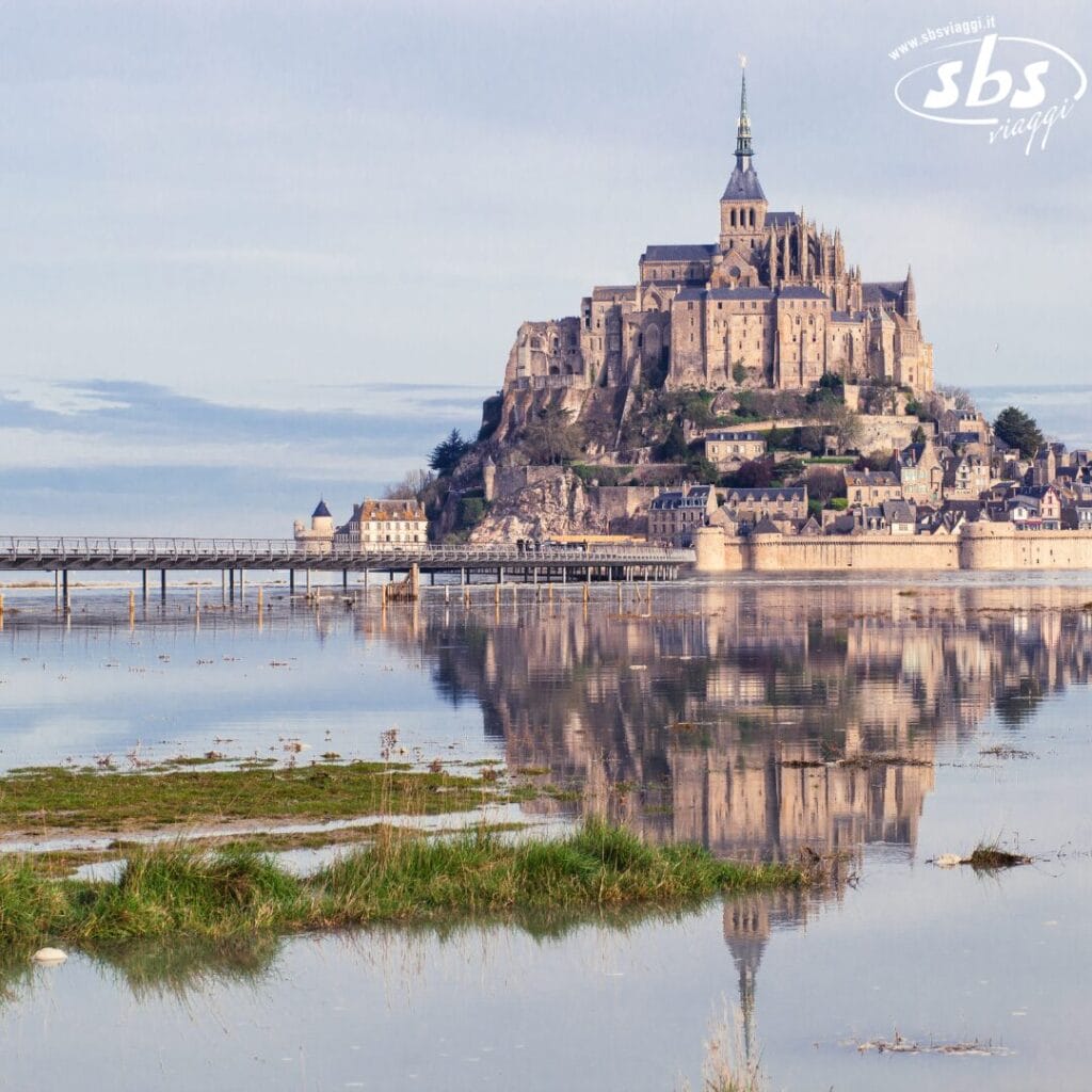 Il Mont Saint-Michel riflesso nell'acqua ferma, con un cielo terso sopra. Le mura fortificate dell'abbazia e il verde creano una scena pittoresca, come se fosse stata realizzata da un artista che creava una bozza automatica di splendore naturale.