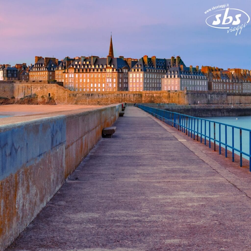 Sentiero costiero che conduce a una città storica cinta da mura, con edifici illuminati e una guglia prominente, sotto un cielo colorato.