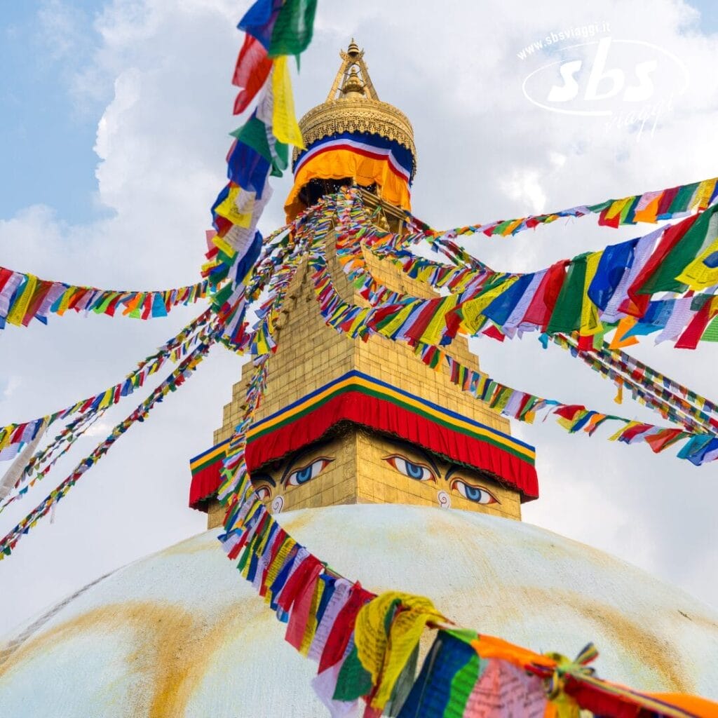 Lo stupa di Boudhanath con le sue colorate bandiere di preghiera e gli occhi dipinti sulla torre, sullo sfondo di un cielo azzurro con nuvole.