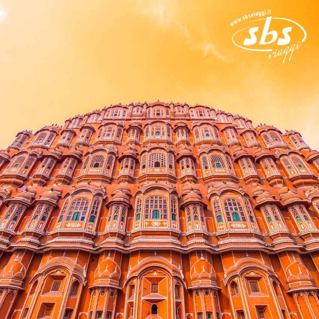 Vista dal basso dell'Hawa Mahal a Jaipur, in India, che mette in mostra la sua intricata facciata in arenaria rossa e rosa sotto un cielo sfumato di arancione; un capolavoro di grandiosità architettonica.