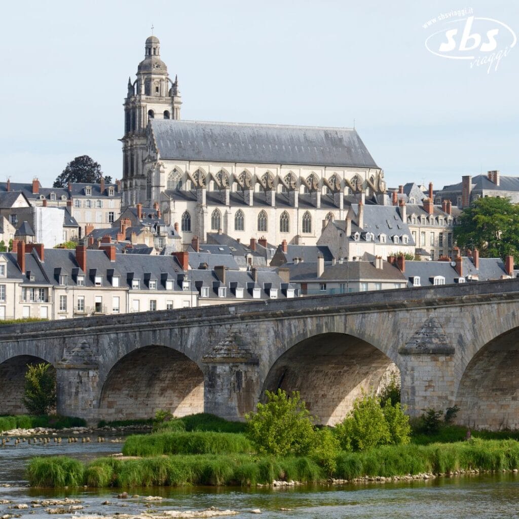 Una cattedrale storica con un alto campanile si erge maestosa dietro un ponte di pietra che attraversa il fiume, circondata da affascinanti edifici cittadini e vegetazione verdeggiante. Bozza automatica cattura questa scena serena in perfetta armonia.