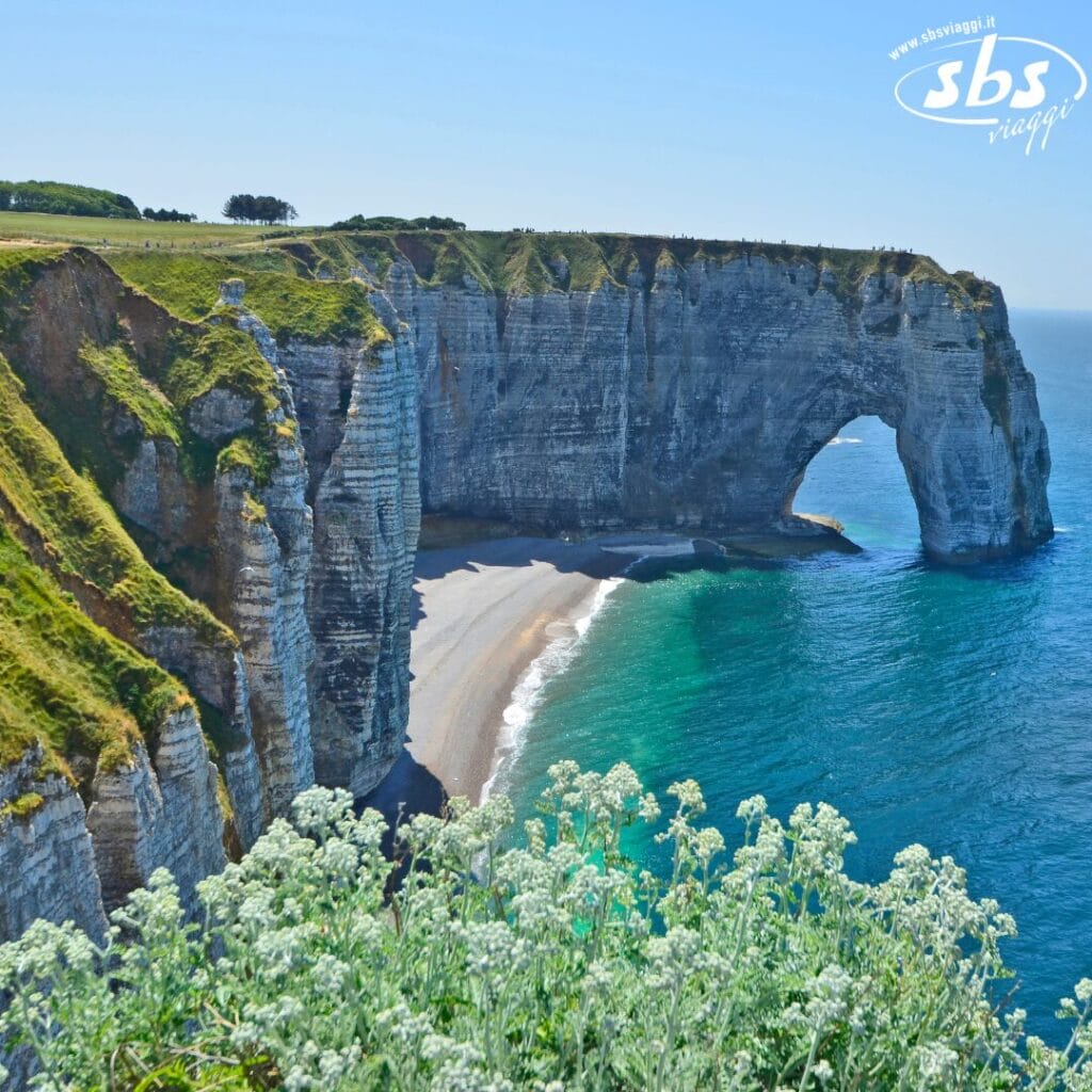 Le scogliere costiere con un arco naturale ornano la spiaggia, incorniciate da verdi colline e un oceano azzurro sotto un cielo limpido, creando uno scenario simile a una bozza automatica di capolavoro della natura.
