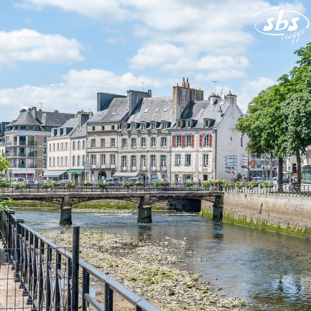 Storico paesaggio urbano europeo con edifici in pietra lungo un fiume, un piccolo ponte e cielo nuvoloso. Ringhiere in ferro sulla riva del fiume e alberi sullo sfondo.