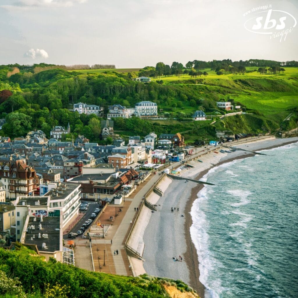 Nella città costiera di Normandia, una spiaggia curva è fiancheggiata da edifici affascinanti e da un lungomare vivace. Verdi colline punteggiate di alberi formano lo sfondo sotto un cielo parzialmente nuvoloso, rendendolo una tappa ideale della tua avventura Gran Tour Parigi.