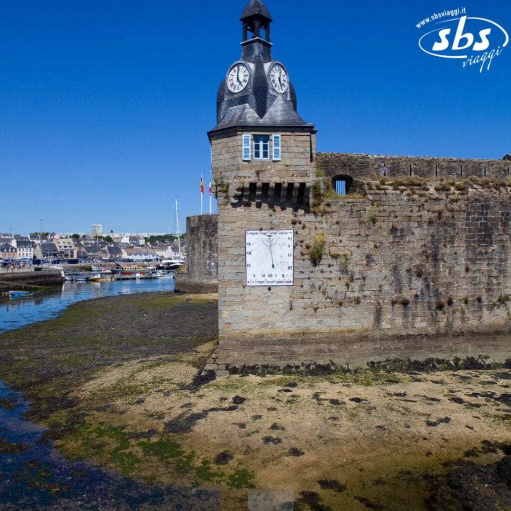 Torre dell'orologio su un muro di pietra di una fortezza vicino all'acqua, con barche ed edifici sullo sfondo sotto un cielo azzurro e limpido.