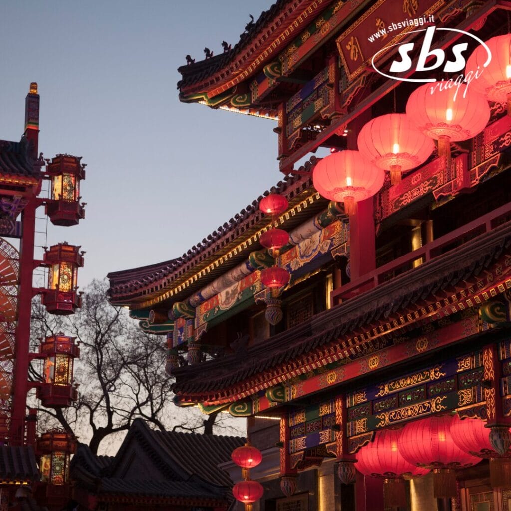 Un edificio ornato con architettura tradizionale cinese è illuminato da lanterne rosse, che proiettano un sublime chiarore durante il tramonto. Alberi e un'altra struttura simile si ergono sullo sfondo, dipingendo una scena incantevole che ricorda l'antica Cina.