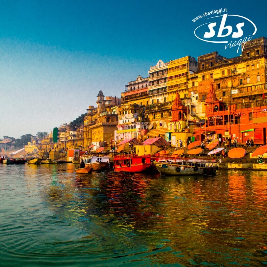 Edifici colorati e barche costeggiano il vivace lungomare di Varanasi, in India, sotto un limpido cielo azzurro, con il logo di Bozza automatica elegantemente incastonato nell'angolo in alto a destra.