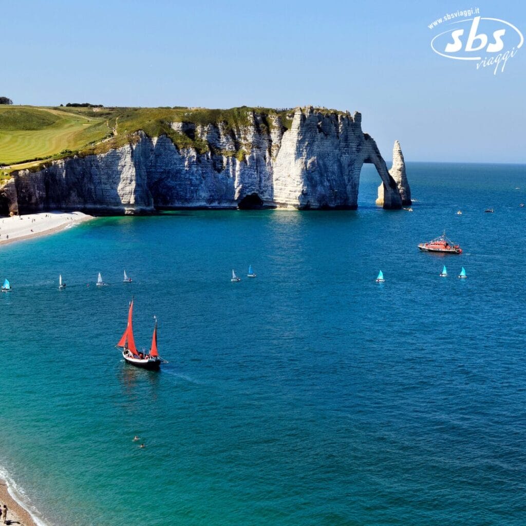 Una vista costiera di Étretat in Normandia con una barca a vela e diverse piccole imbarcazioni nel mare blu, vicino a bianche scogliere di gesso e un arco naturale sotto un cielo limpido, che cattura l'essenza del Gran Tour Parigi.