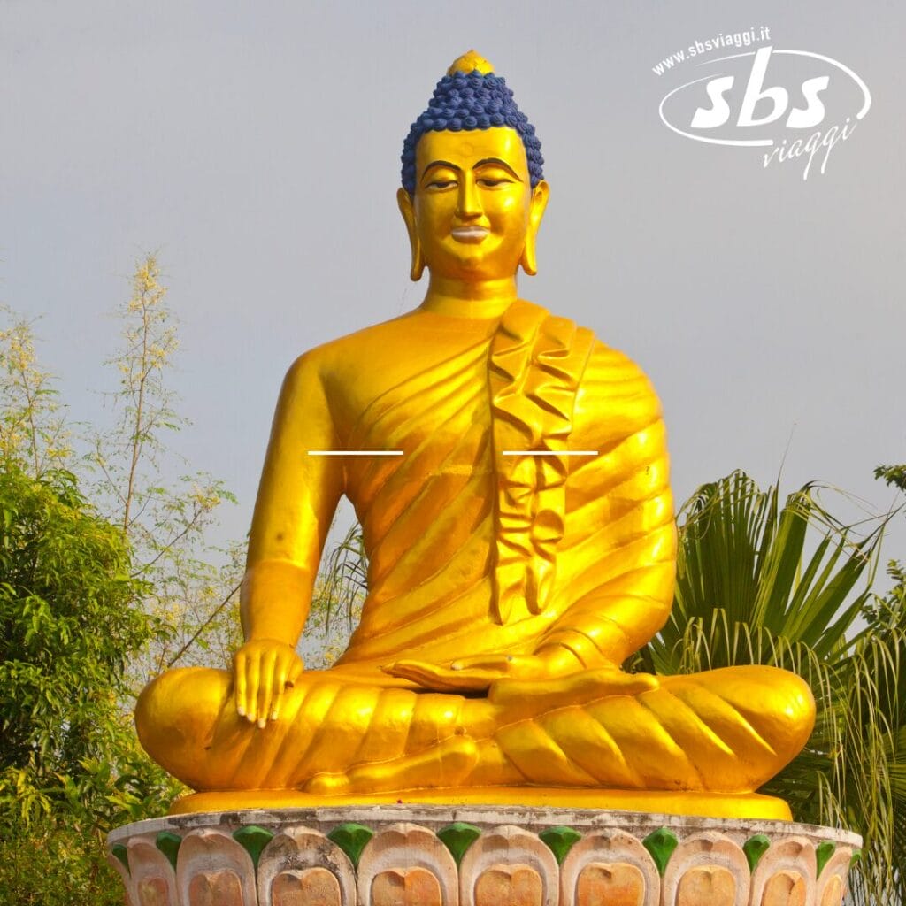 Statua dorata del Buddha seduto a gambe incrociate su un piedistallo di loto, con alberi sullo sfondo e il logo "sbs viaggi" in alto a destra.