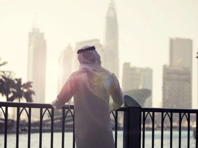 Vestito con abiti tradizionali, il soggetto si appoggia casualmente a una ringhiera, ammirando lo skyline della città con i suoi imponenti edifici. La scena sembra quasi una "bozza automatica", che cattura senza sforzo un momento urbano ricco di echi culturali.