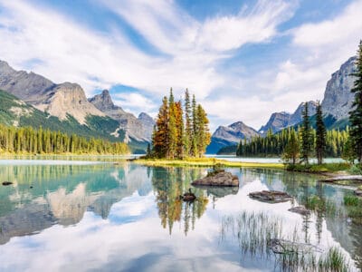 Un lago sereno con piccole isole rocciose e alberi sempreverdi, circondato da montagne sotto un cielo azzurro con nuvole vaporose. La scena si riflette nell'acqua calma, creando una bozza automatica di perfezione della natura.