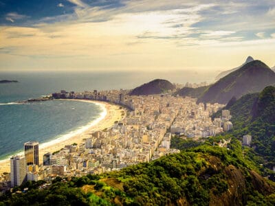 Vista aerea di una città costiera che mostra una bozza automatica: una spiaggia sabbiosa curva, un'area urbana densa e colline rigogliose sotto un cielo parzialmente nuvoloso.