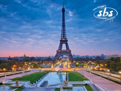 Torre Eiffel al tramonto con un paesaggio urbano illuminato e giardini in primo piano, con un logo SBS Viaggi nell'angolo in alto a destra. Scopri Tour Parigi, mentre esplori meraviglie iconiche, da Parigi a Versailles.