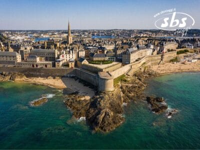 Vista aerea di una fortezza costiera con spiagge sabbiose, costa rocciosa ed edifici storici. Paesaggio urbano con una guglia di chiesa prominente sullo sfondo.