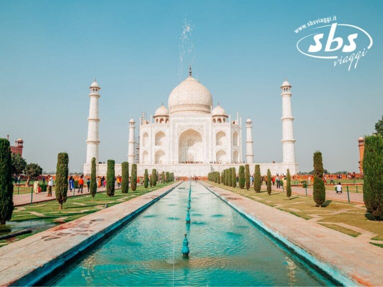 Il Taj Mahal ad Agra, in India, è splendidamente ripreso frontalmente, con piscine serene e riflettenti e giardini lussureggianti che ne esaltano la maestosità. Nell'angolo in alto a destra, un logo "sbs viaggi" completa sottilmente la scena con eleganza italiana.