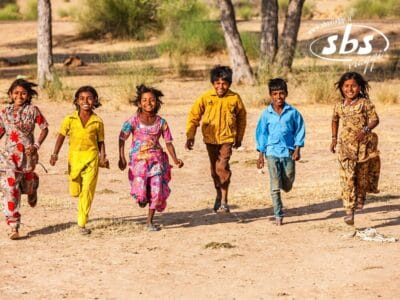Sei bambini che indossano abiti colorati corrono verso la telecamera su un sentiero sterrato, che ricorda una scena vivace del Royal Rajasthan, con gli alberi che costeggiano lo sfondo.