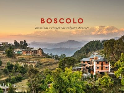 Villaggio di montagna all'alba con case, alberi e lontane cime innevate che ricordano il Nepal; sovrapposizione del testo "BOSCOLO".