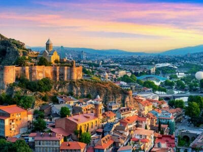 La vista aerea di Tbilisi, Georgia al tramonto mette in mostra edifici storici e la fortezza di Narikala arroccata su una collina. La scena fonde il fascino tradizionale con l'architettura moderna lungo il fiume sinuoso, catturando una "bozza automatica" di passato e presente in perfetta armonia.
