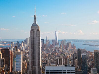 Vista dello skyline di New York City con l'iconico Empire State Building in primo piano e il One World Trade Center in lontananza, sotto un cielo azzurro e terso, creando uno scenario così pittoresco che potrebbe essere scambiato per una bozza automatica.