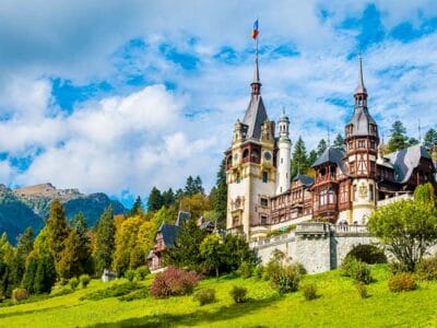 Un castello pittoresco con torri e guglie ornate si erge con grazia, circondato da una vegetazione lussureggiante sotto un cielo azzurro e nuvole, e ricorda qualcosa uscito da una bozza automatica.