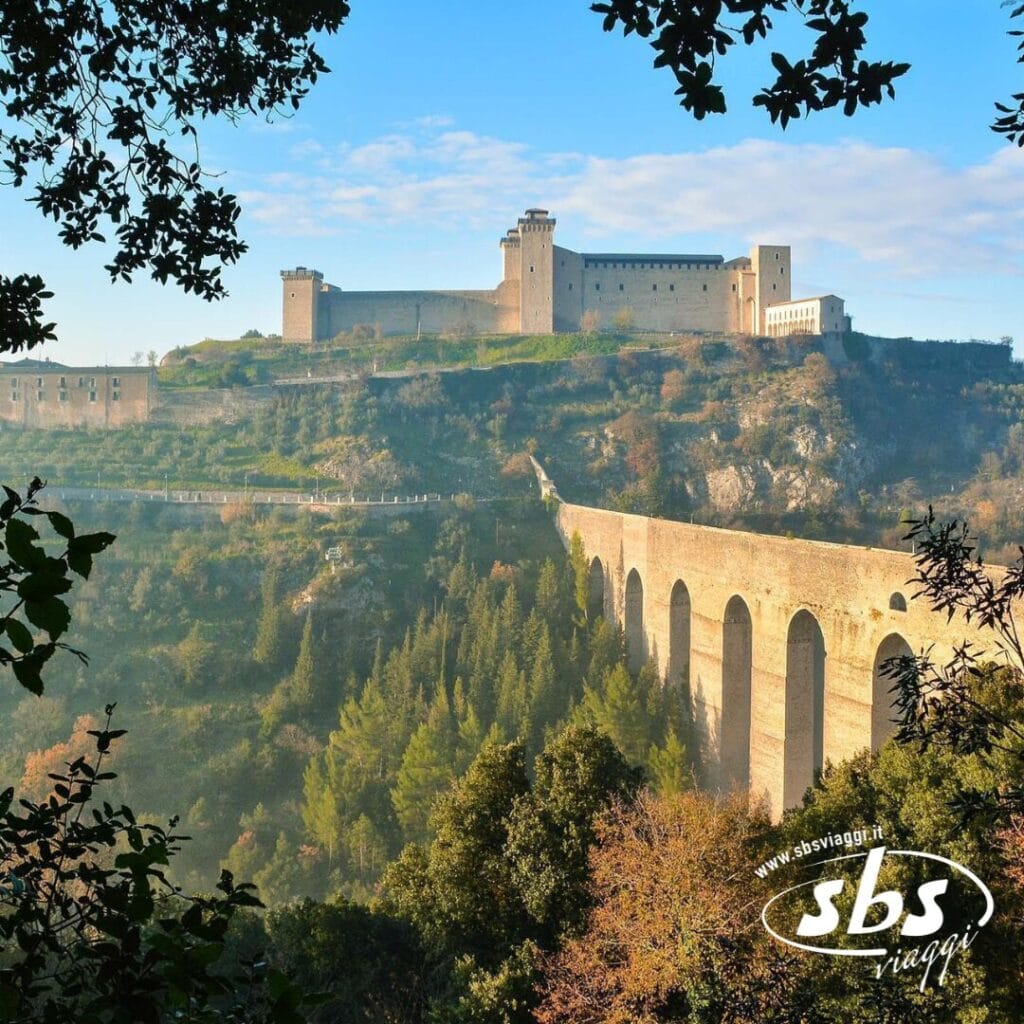 Situata sulla cima di una collina in Umbria, questa fortezza medievale vanta uno straordinario acquedotto in pietra e una vegetazione lussureggiante in primo piano, il tutto sotto un cielo azzurro e terso, rendendola una meta turistica perfetta.