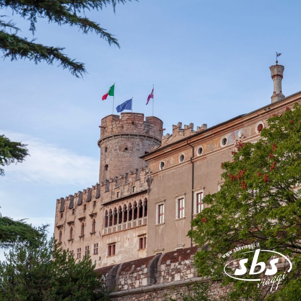 Un maestoso castello in pietra con una torre rotonda, ornato da bandiere italiane, si erge contro il cielo azzurro. Il verde incornicia la scena in Trentino Alto Adige.